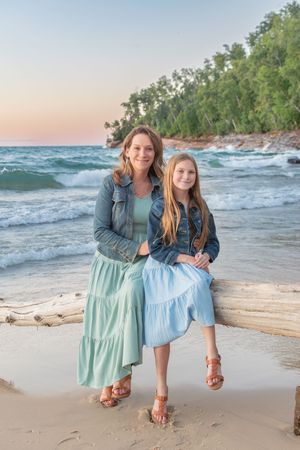 Two people in coordinating blue and mint dresses pose playfully on a beach at sunset with waves crashing behind them.