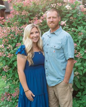 A couple poses together in front of pink flowering bushes, with her wearing a blue dress and him in a light denim shirt.