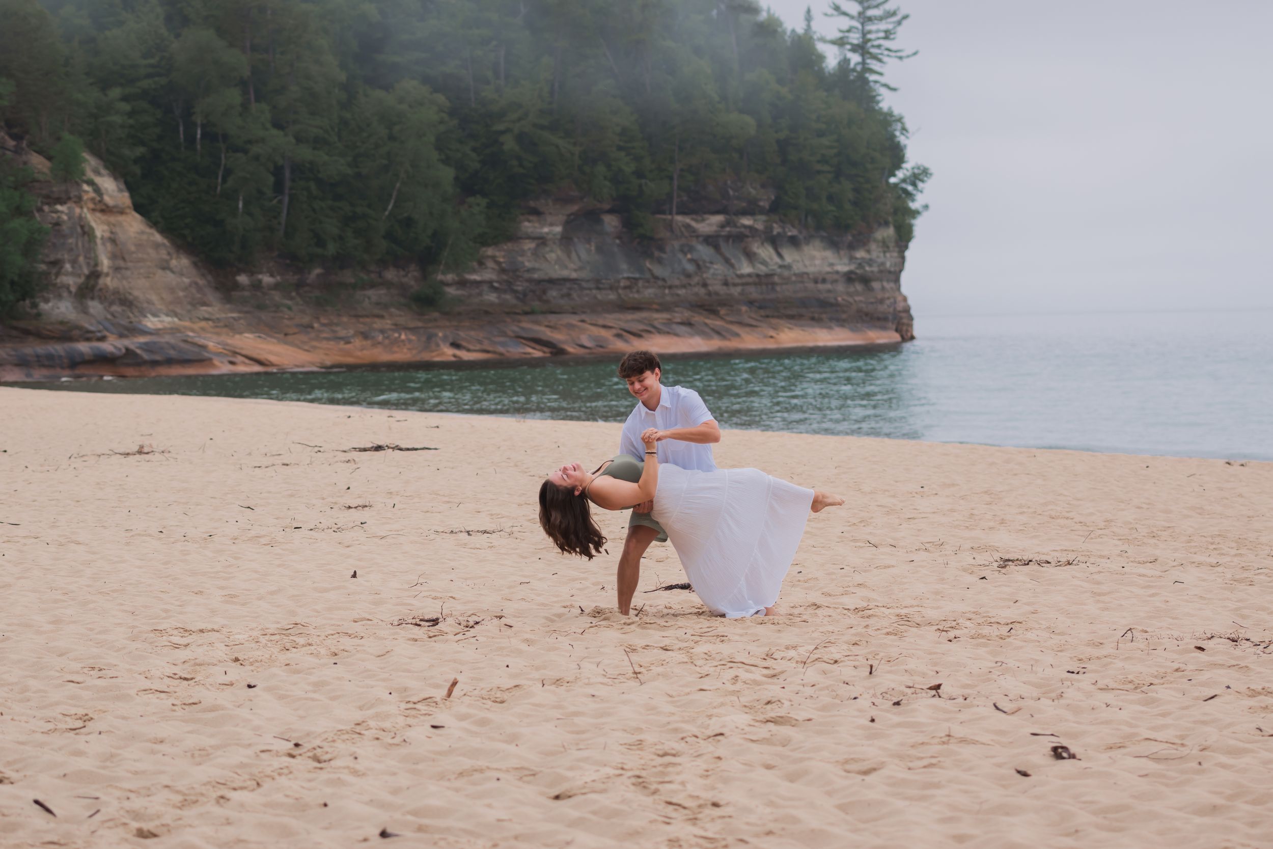 A couple poses together on a scenic sandy beach with rocky cliffs and ocean views in the background.