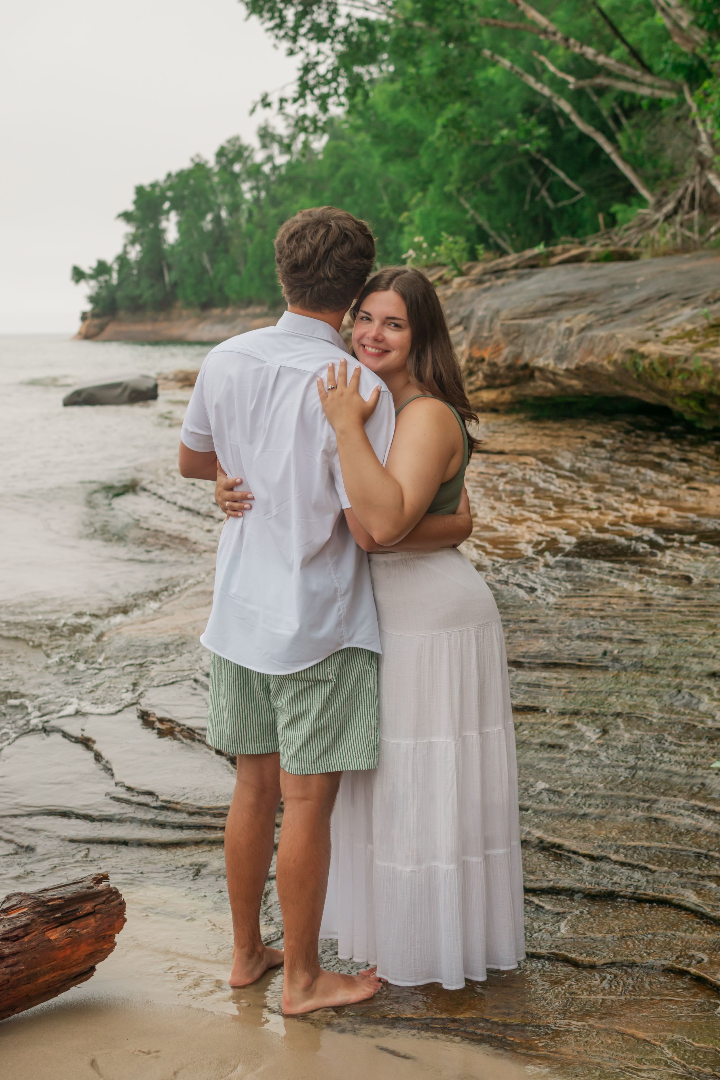 A couple shares a romantic moment on a rocky beach with lush green trees in the background.