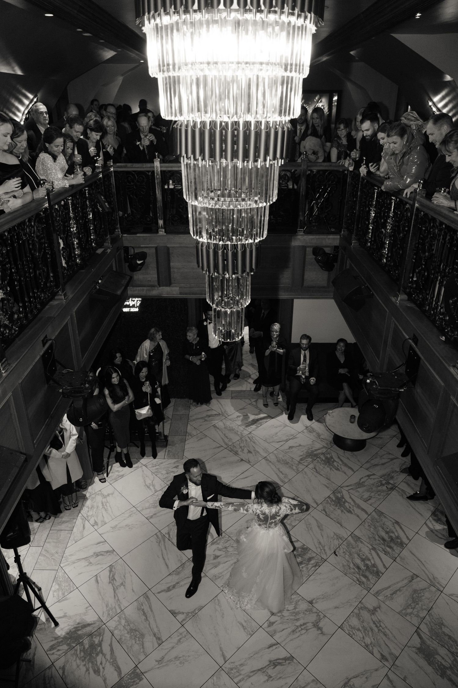 Black and white photo of a couple having one of their first dances with all their guests watching from above and below