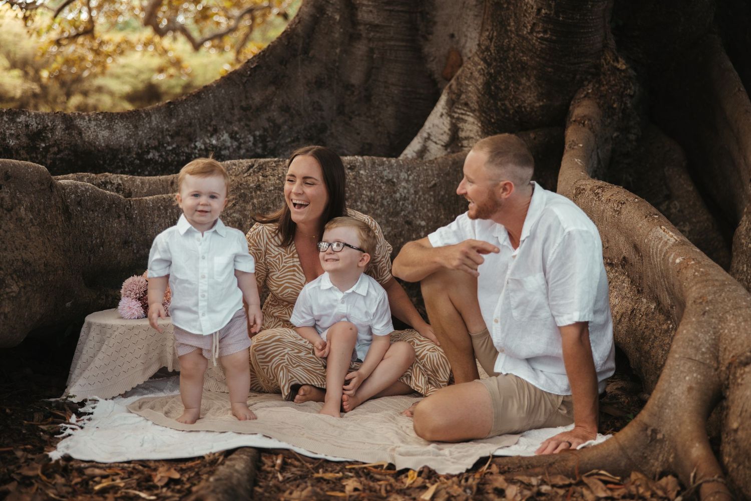 A small family sits together on a blanket between large tree roots at sunset in an outdoor setting.