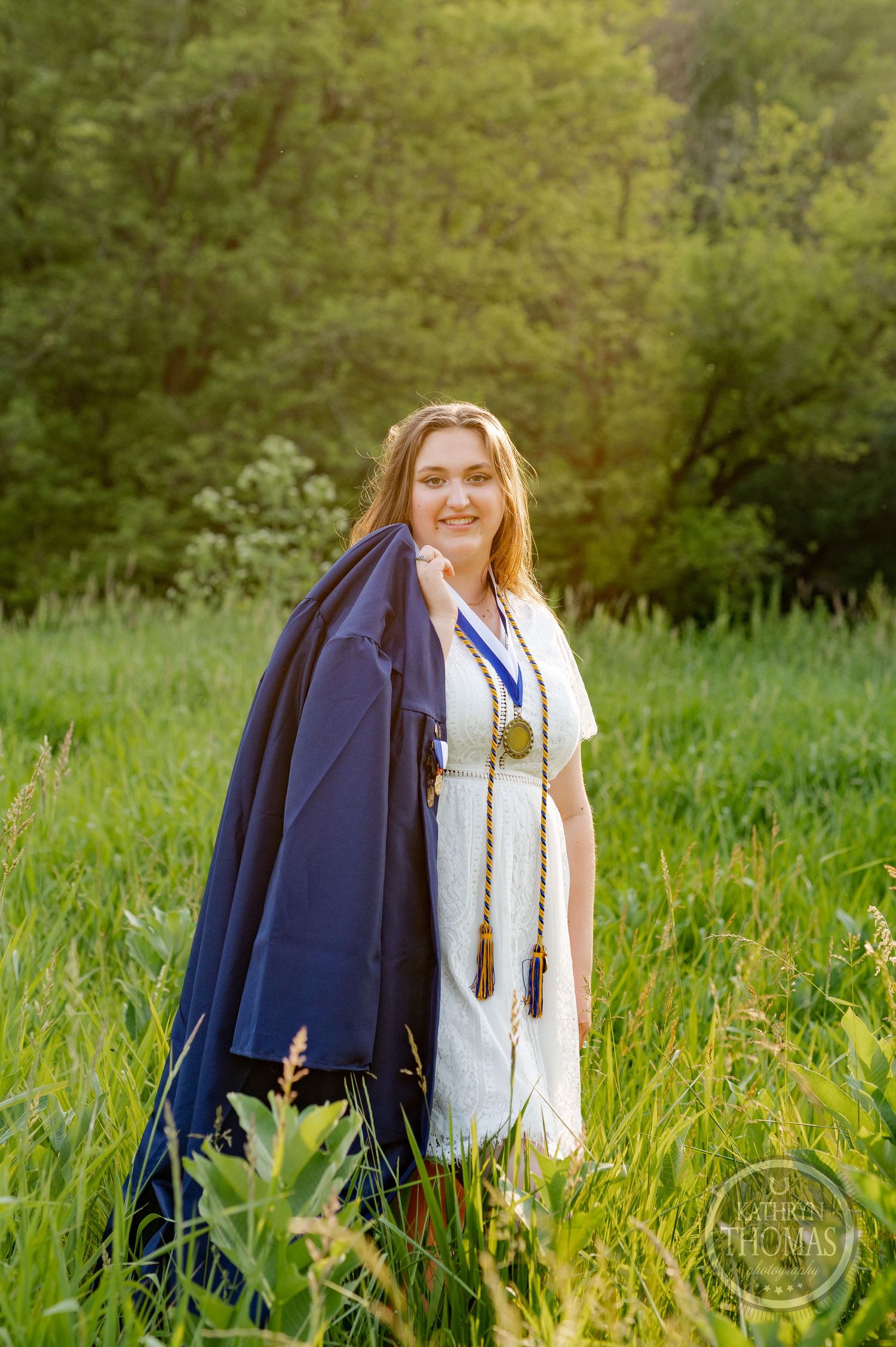 Graduate in white dress and navy blue cape standing in sunlit grassy field.