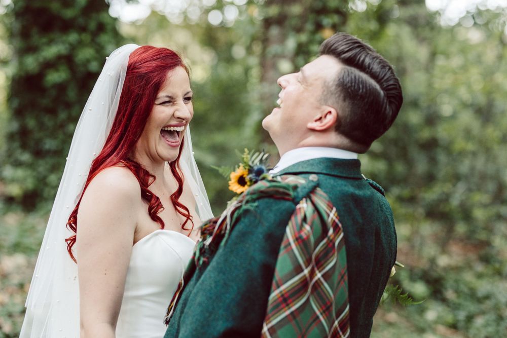 Wedding couple in Scottish attire laughing together in a sunlit forest.