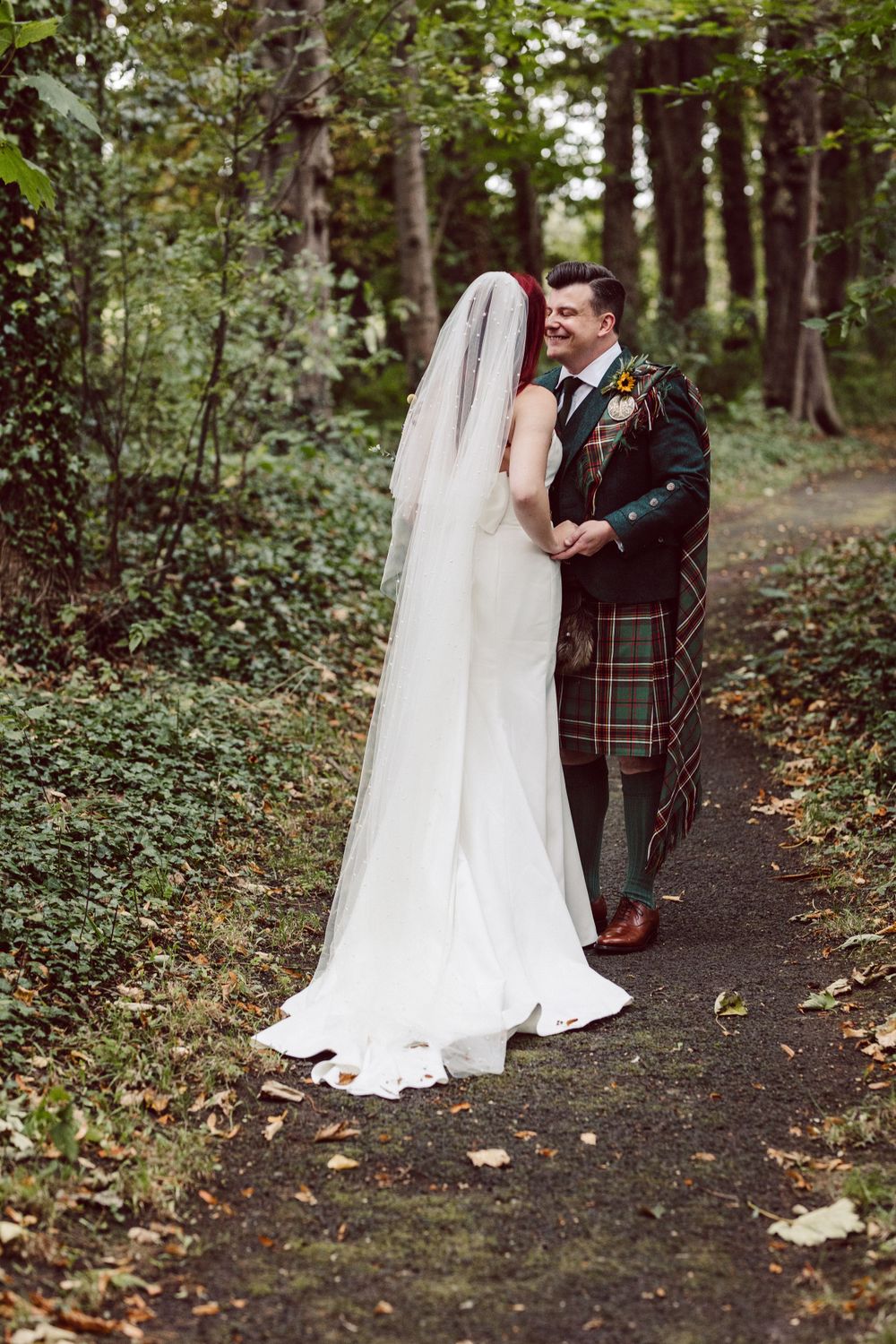 A bride and groom in Scottish wedding attire stand on a forest path during their wedding photoshoot.