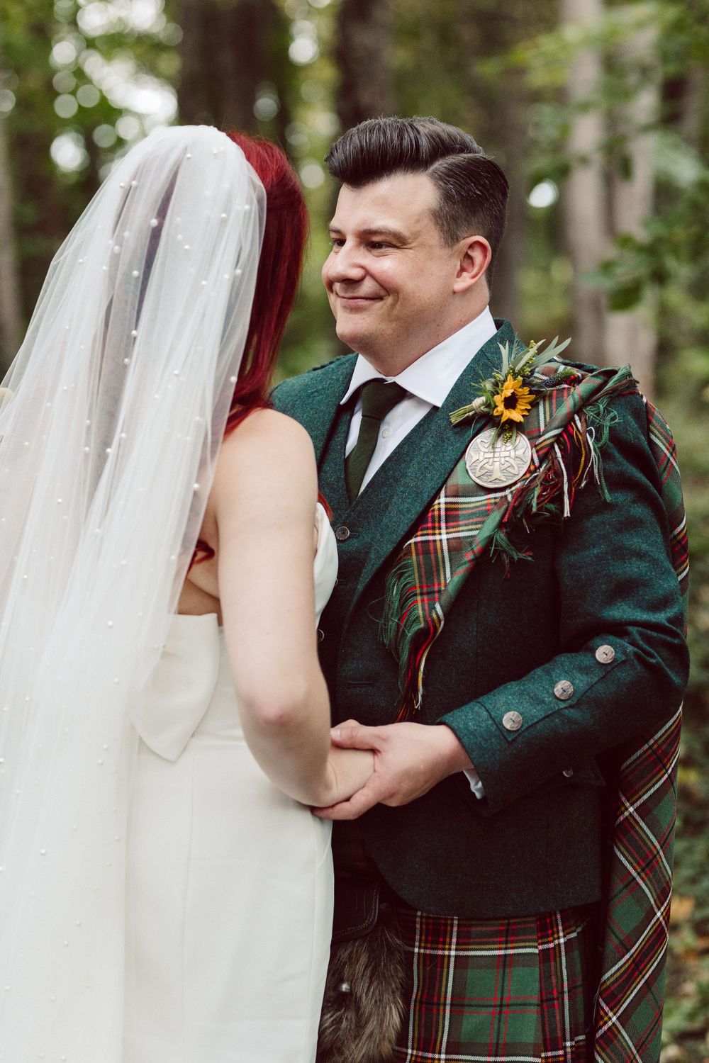 A wedding couple shares an intimate moment in a forest setting with the groom wearing a traditional green Scottish kilt.