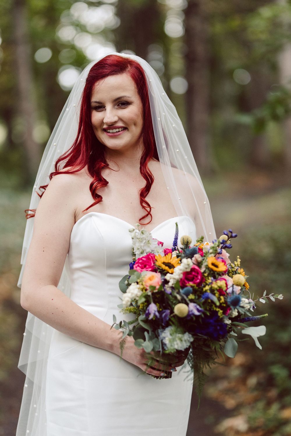 A bride in a white dress holds a vibrant multicolored bouquet against a wooded backdrop.