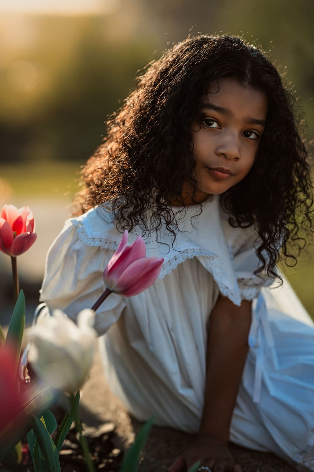 young girl with brown curly hair and white dress sits next to tulips