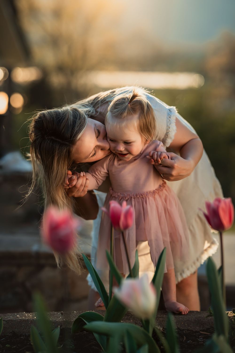 mother leans over her young daughter and kisses her cheek as she helps her stand behind tulips