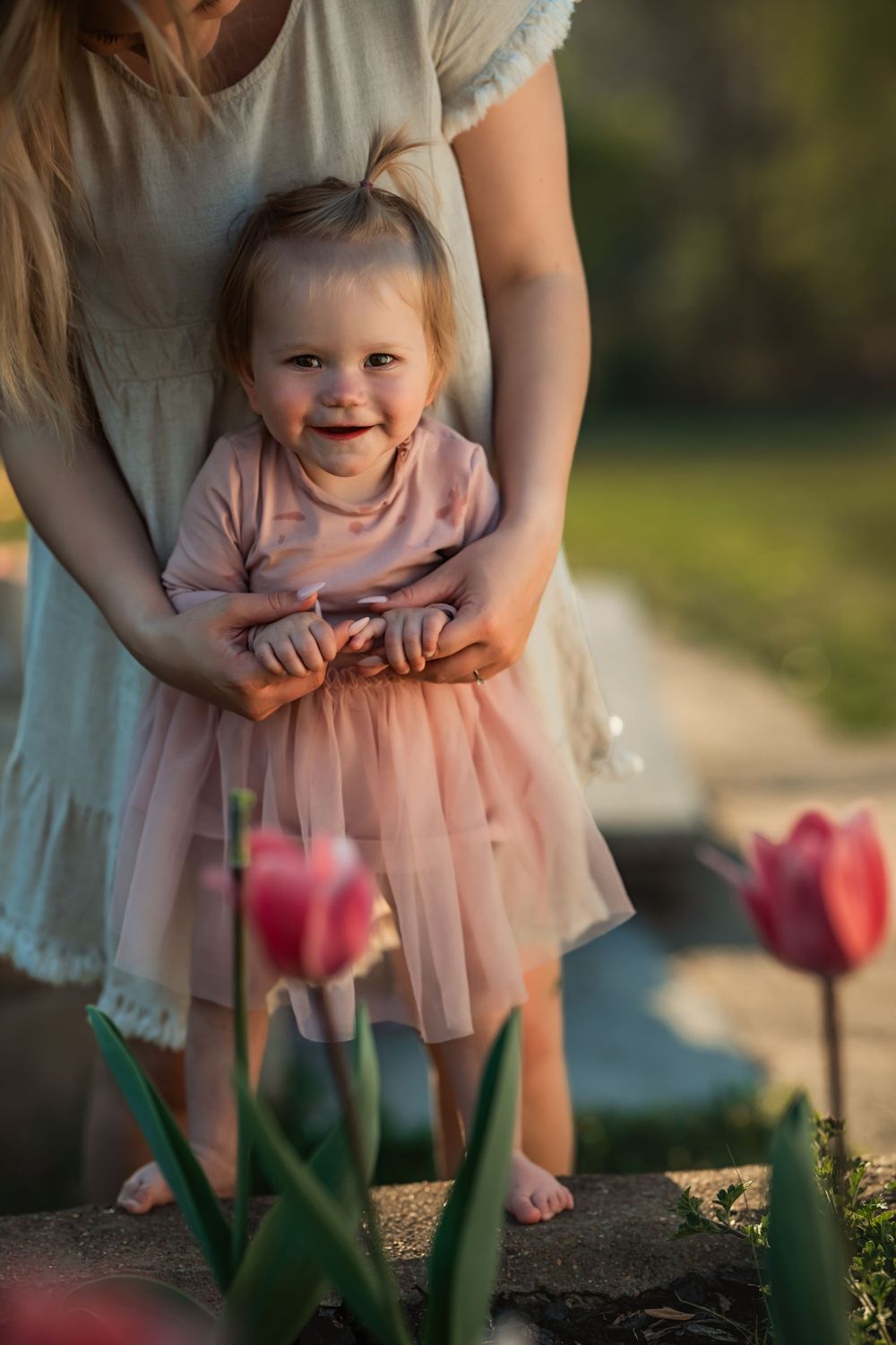 baby girl smiles as her mom helps her stand next to some tulips