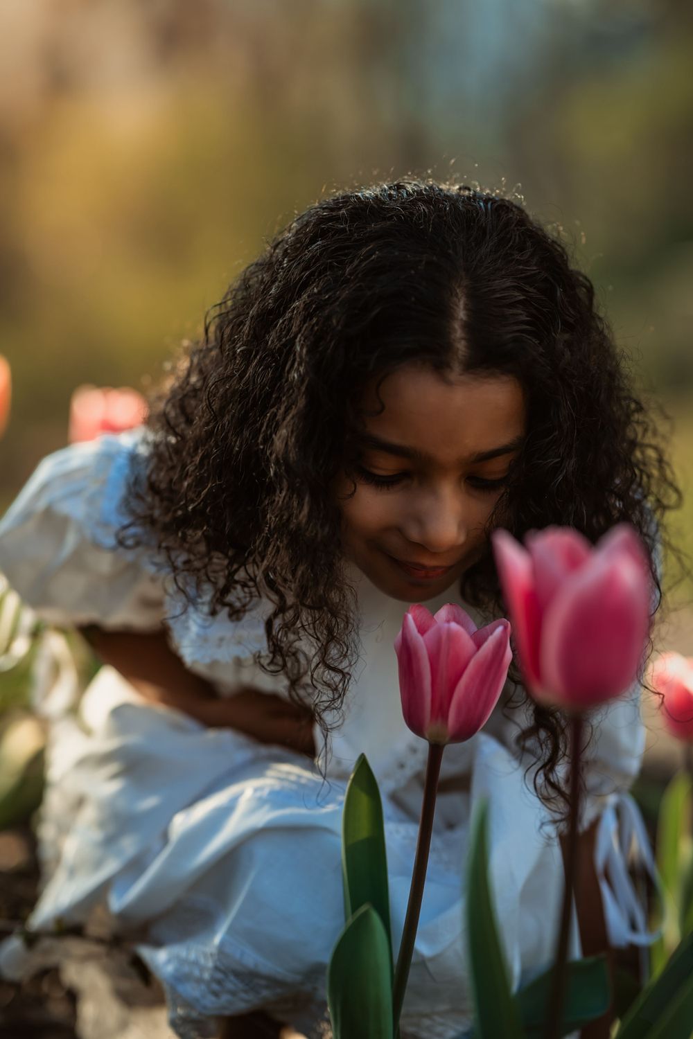 young girl in white dress with brown skin leans over to smell a pink tulip
