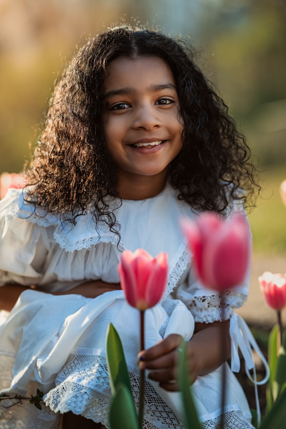young indian girl in white dress crouches behind some tulips and smiles