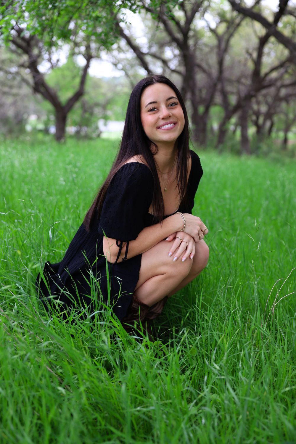 A person in a black dress sits crouched in a lush green grassy field surrounded by trees.