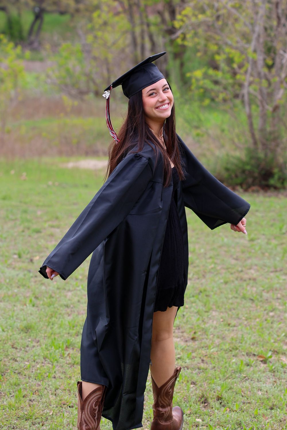A graduate in cap, gown and cowboy boots poses playfully in a grassy field.