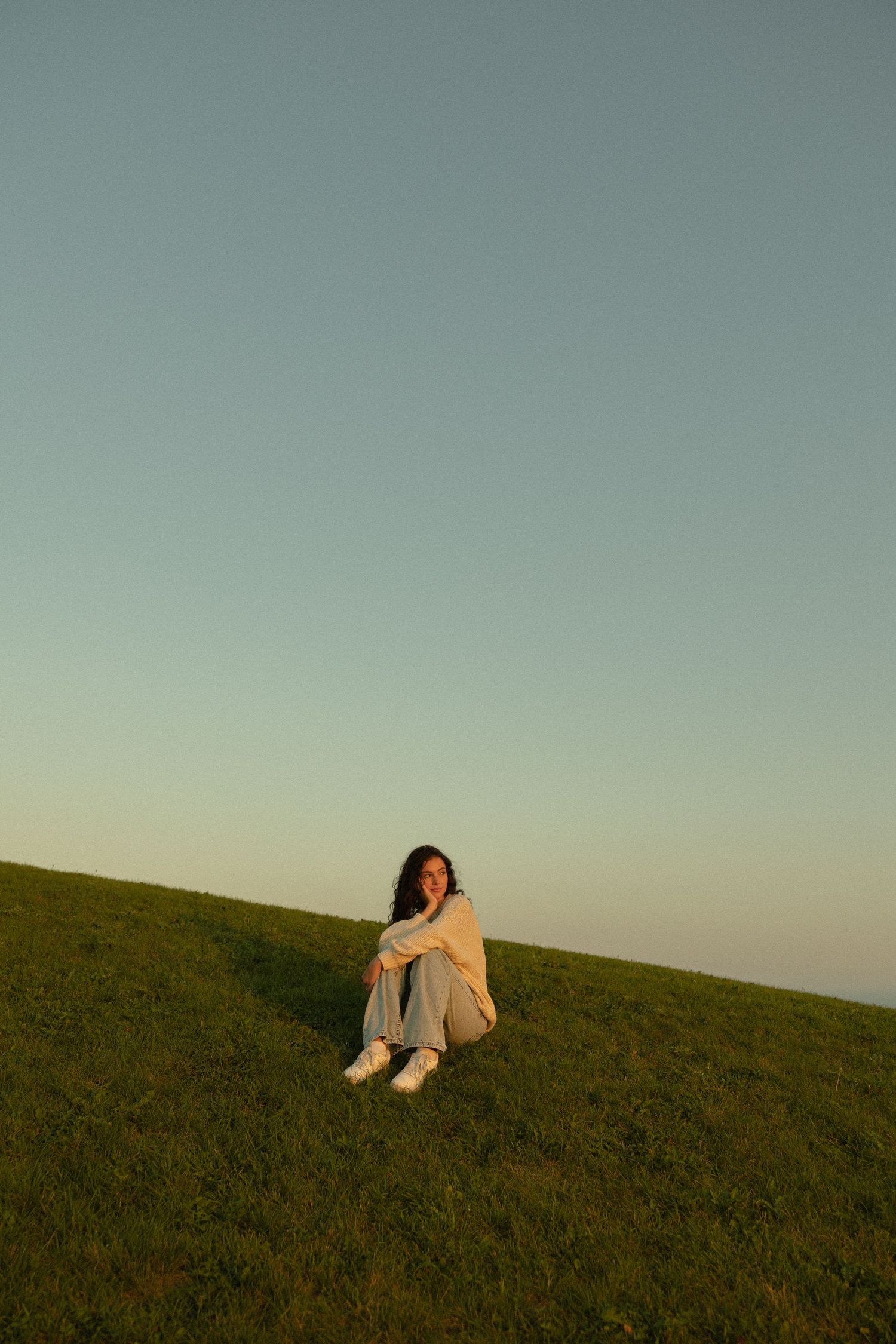 A solitary figure sits on a grassy hillside during golden hour.