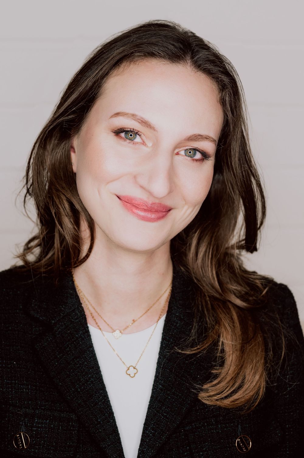 Professional headshot of a woman wearing a black blazer and layered necklaces against a light background.