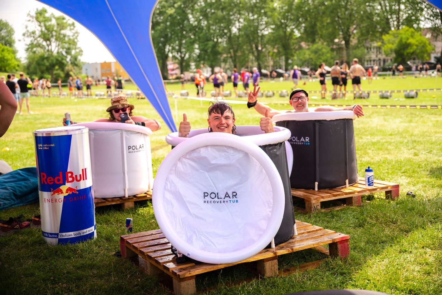 People enjoying drinks and coolers on wooden pallets at an outdoor summer festival in a grassy park setting.