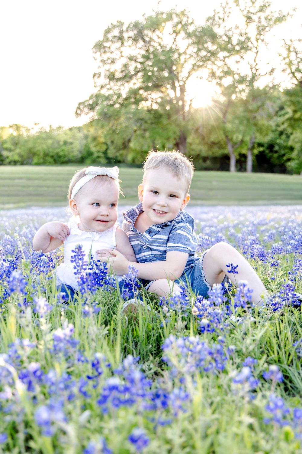 Two young children sit among vibrant purple bluebonnets in a sunny field during golden hour.