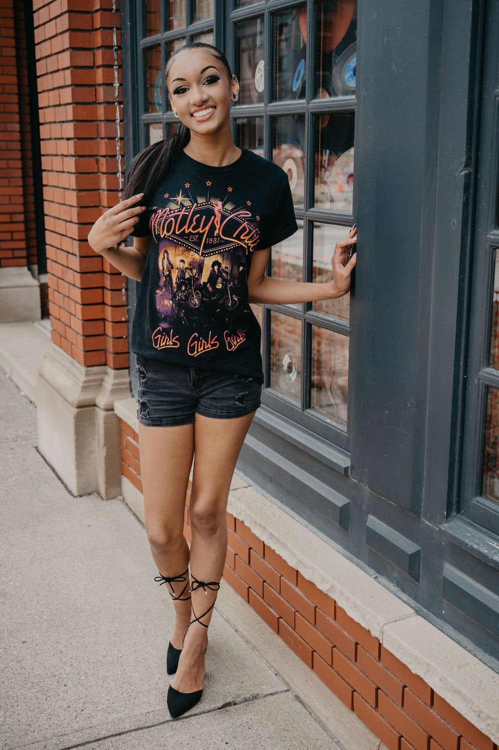 Smiling woman in Motley Crue tee and denim shorts poses against a brick building in black heels.