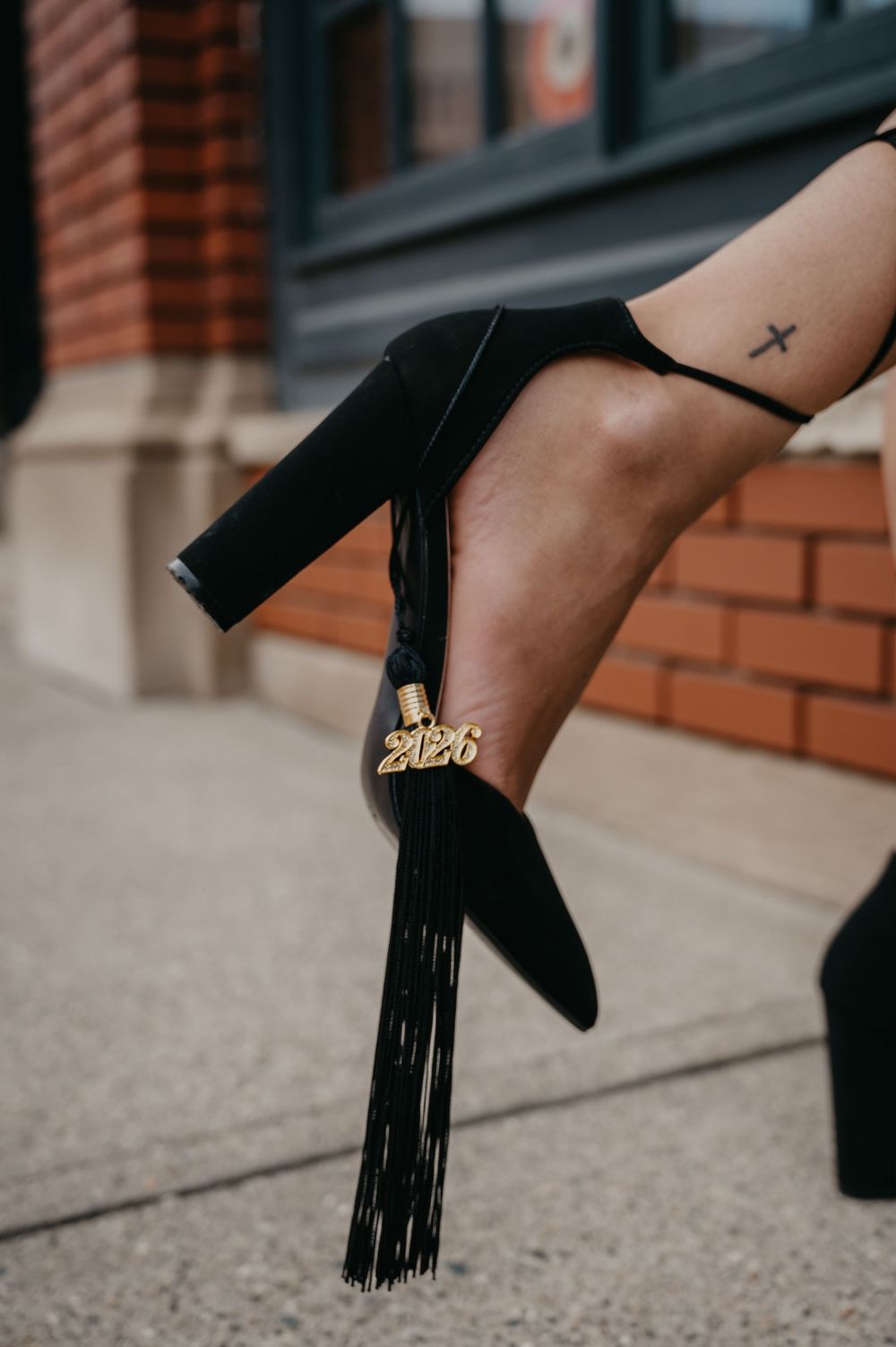Close-up of a black suede fringe heel with gold 2026 charm and a small cross ankle tattoo.