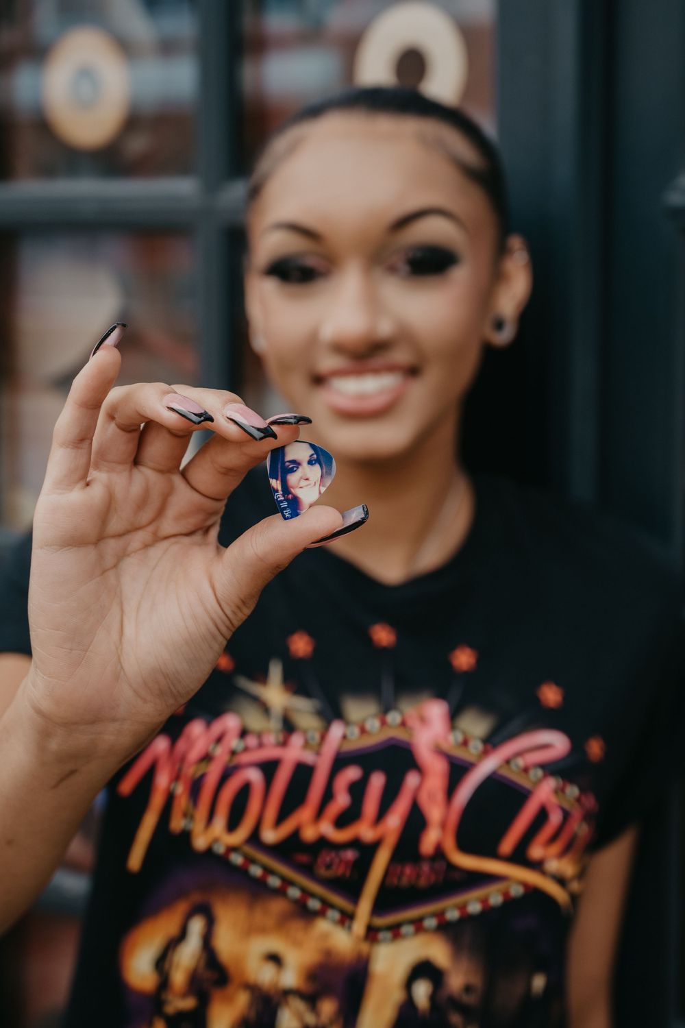Smiling woman in Motley Crue shirt holds up a small decorative pin in front of a storefront.