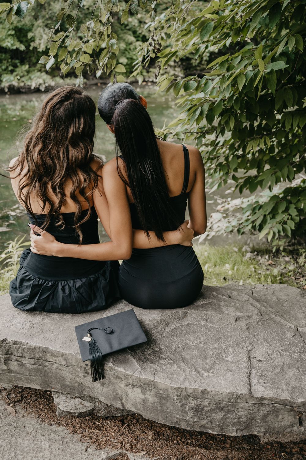 Two young women in black outfits sit embracing on stone ledge with graduation cap beside them.
