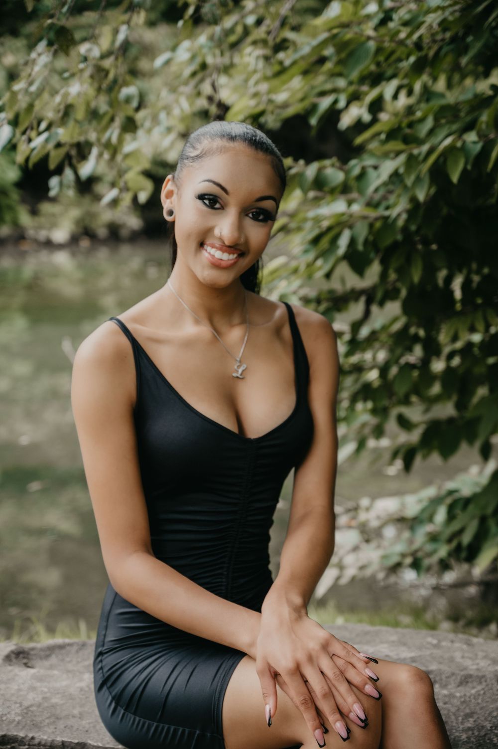 Smiling young woman in black fitted dress sits on stone ledge surrounded by lush green foliage.