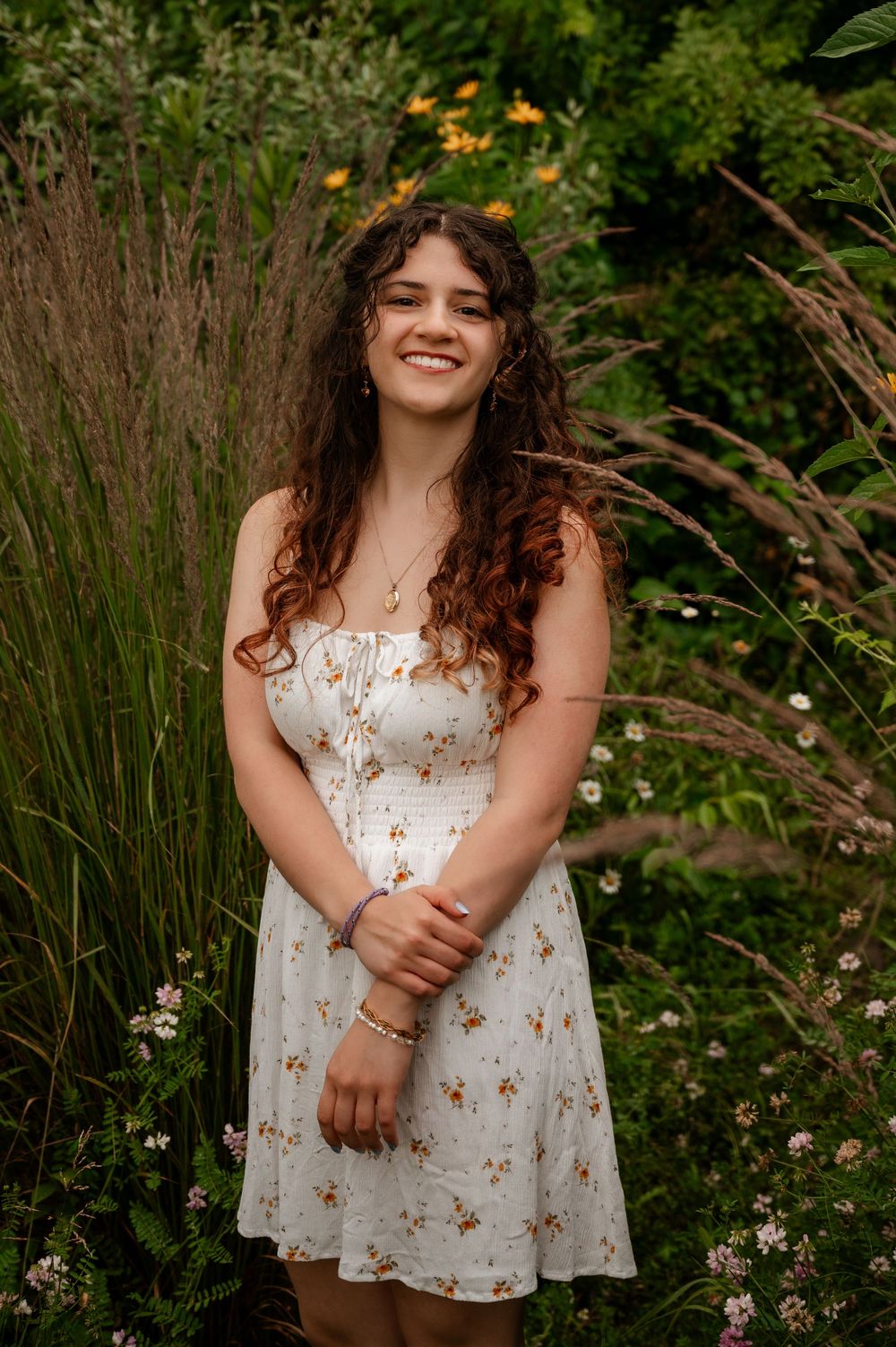 Cheerful young woman in white floral dress standing in lush garden for senior portraits.