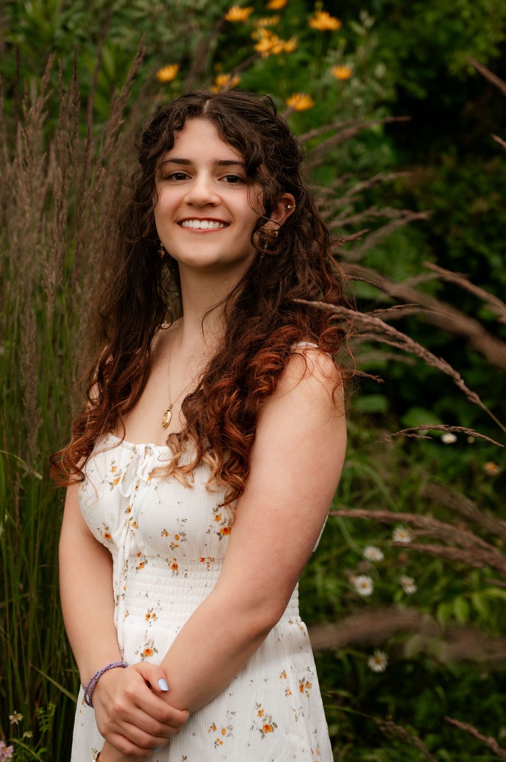 Smiling young woman in floral sundress posing among tall grasses and wildflowers outdoors.