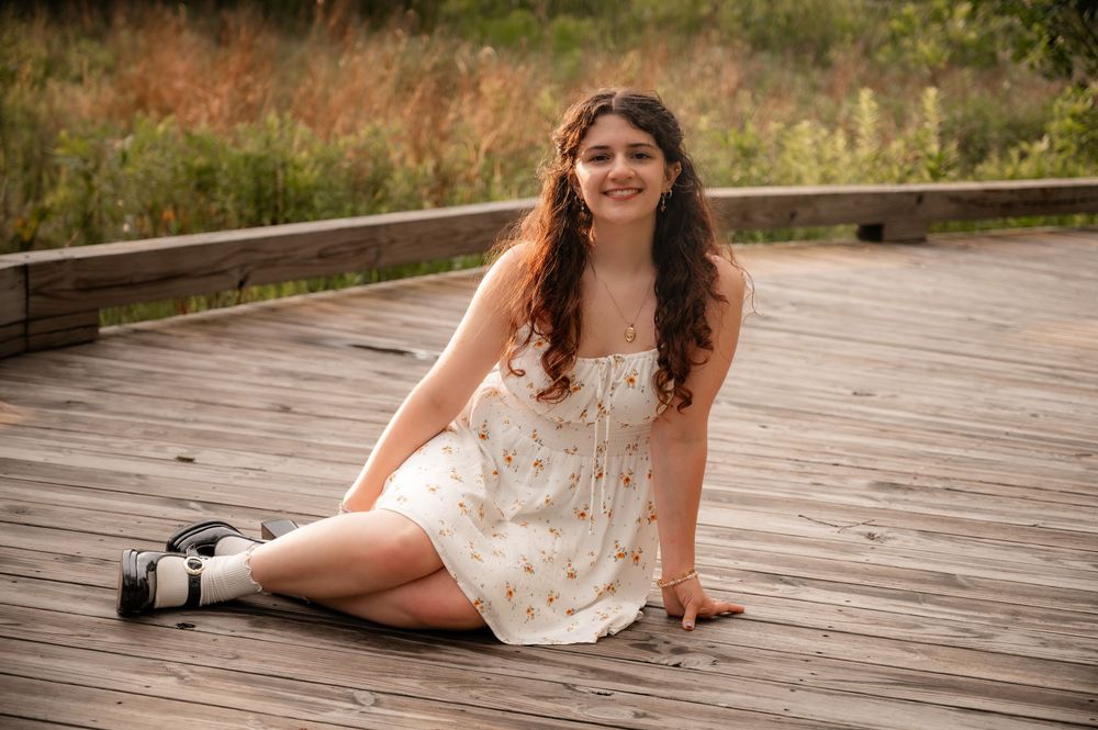 Smiling young woman in floral dress sitting on wooden boardwalk for senior photos.