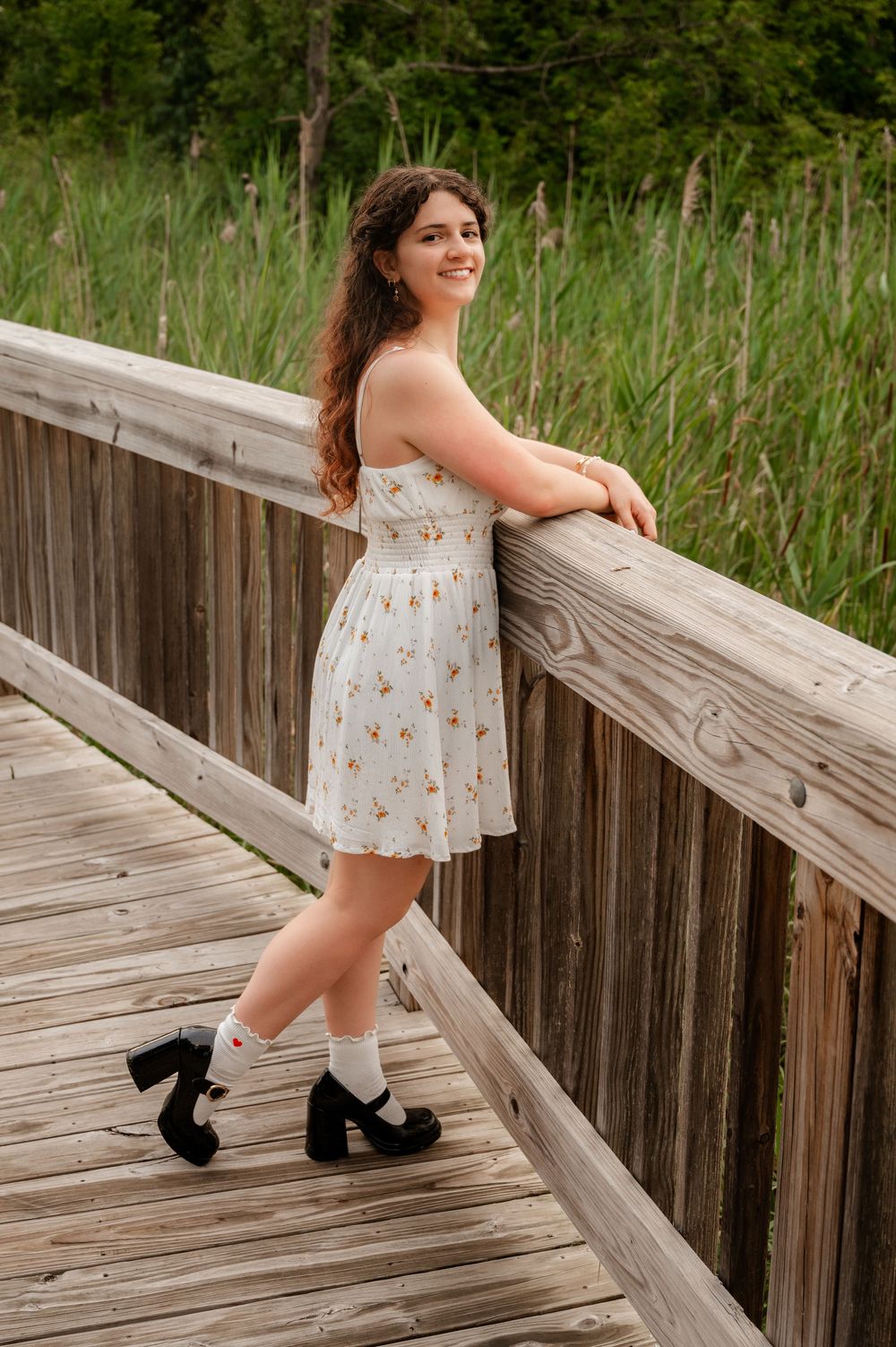 Young woman in white floral dress leaning on wooden bridge railing for senior portraits.
