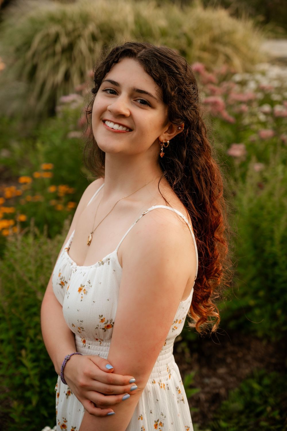 Smiling young woman with curly hair wearing a floral sundress poses in a garden with colorful blooms.
