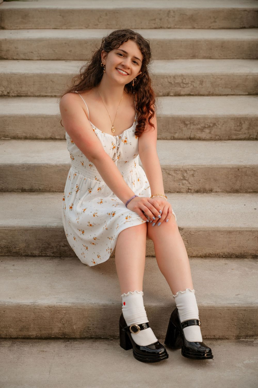 Smiling young woman in floral dress sitting on stone steps wearing black Mary Jane shoes.