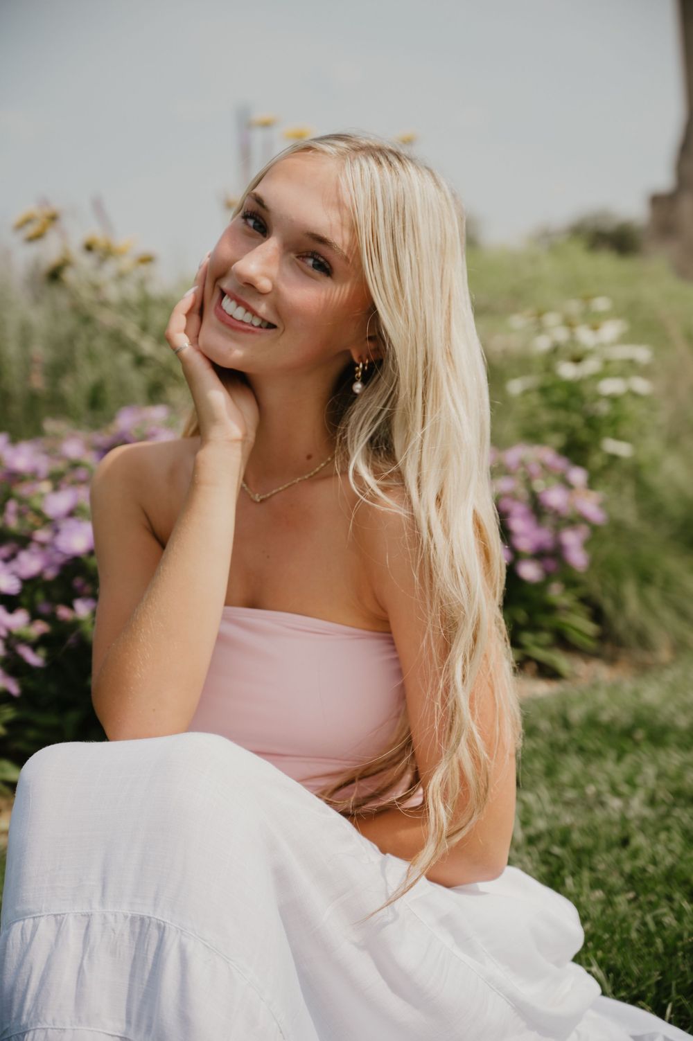 Smiling blonde woman in pink tube top and white skirt sitting among colorful wildflowers outdoors.