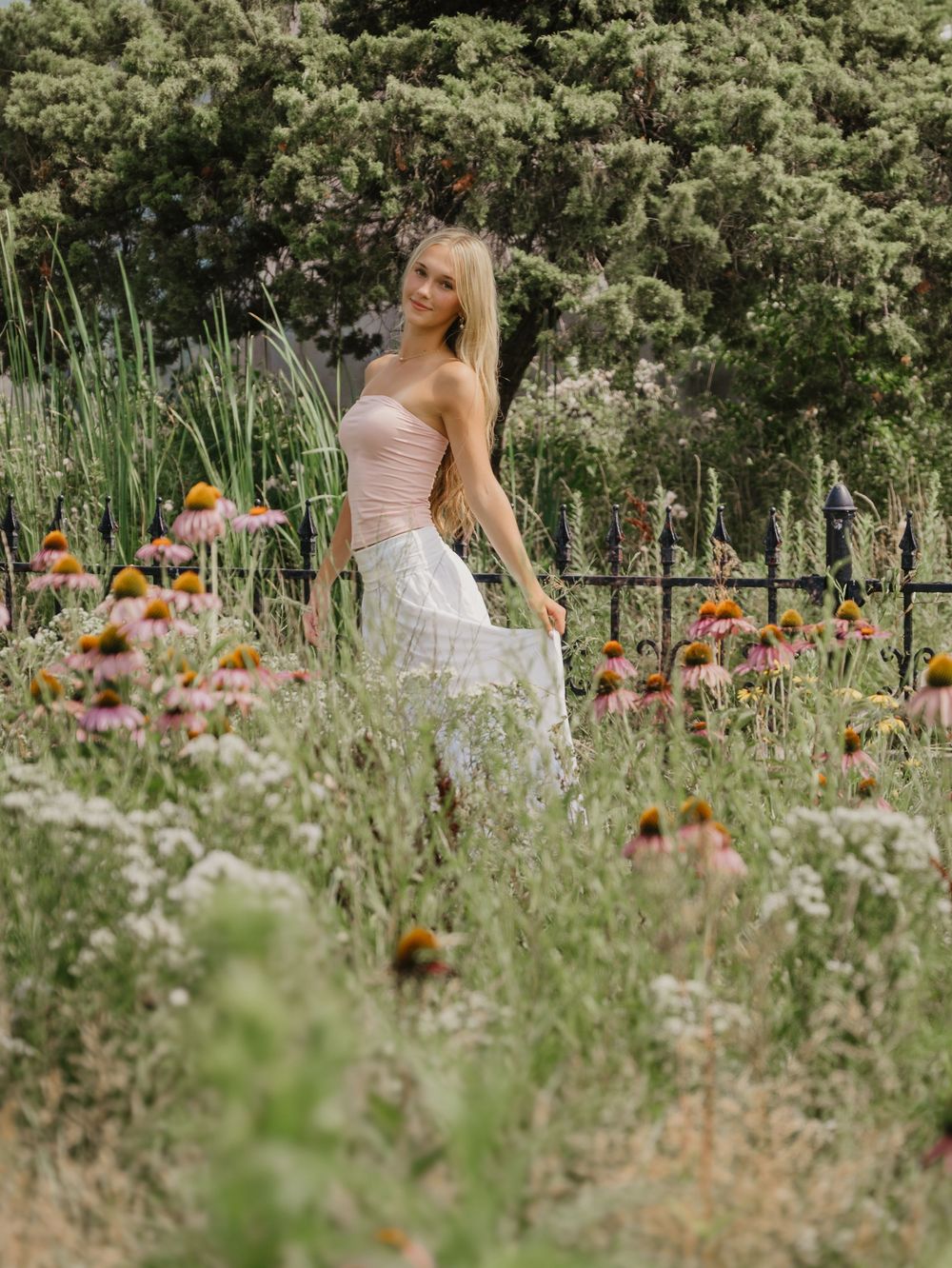 A young blonde woman in a pink top and white skirt stands among wildflowers and coneflowers in a lush garden.