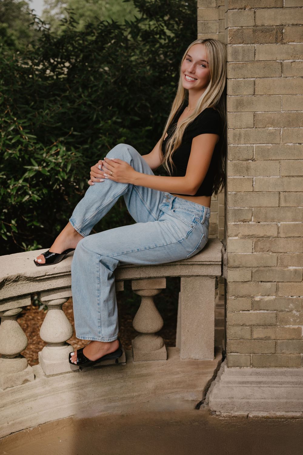 Young blonde woman smiling, sitting on stone balustrade in black crop top, light jeans, and black sandals.