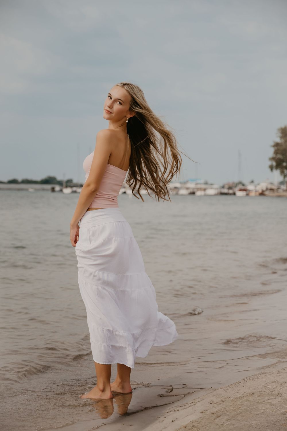 Woman in white skirt and pink top posing on sandy beach with hair blowing, boats visible in background.
