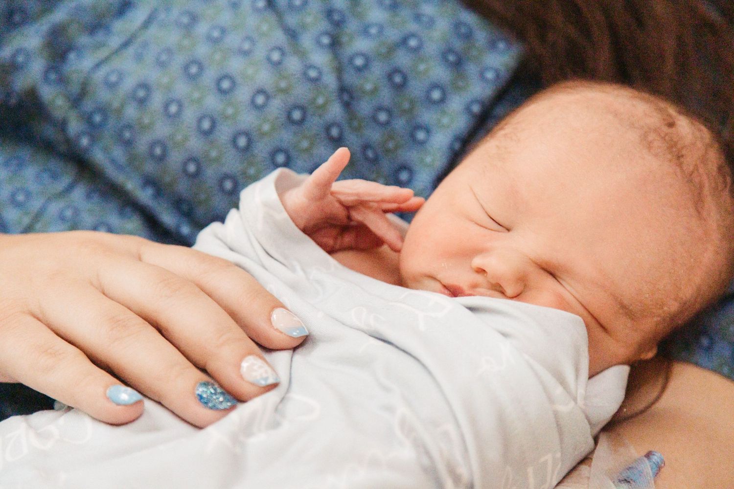 A sleeping newborn baby in white clothing rests peacefully against a blue patterned background.