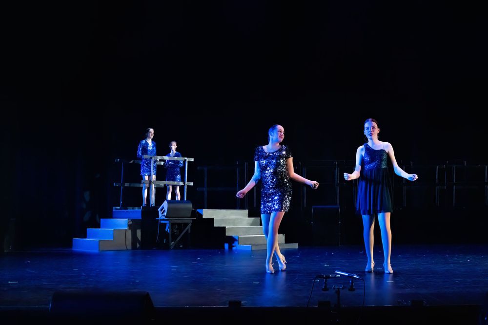 Three performers in blue costumes dance on a dark stage with steps.
