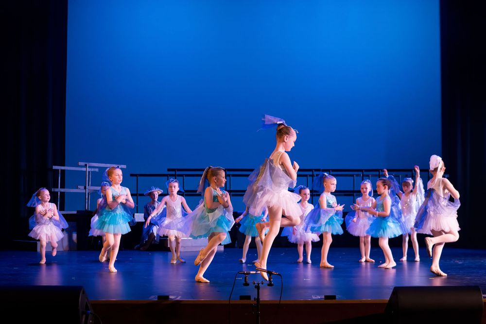 Ballet dancers in light blue tutus perform a synchronized routine on stage against a deep blue backdrop.