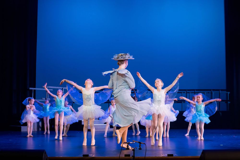 Ballet performance featuring dancers in blue tutus with a central figure in a period costume.