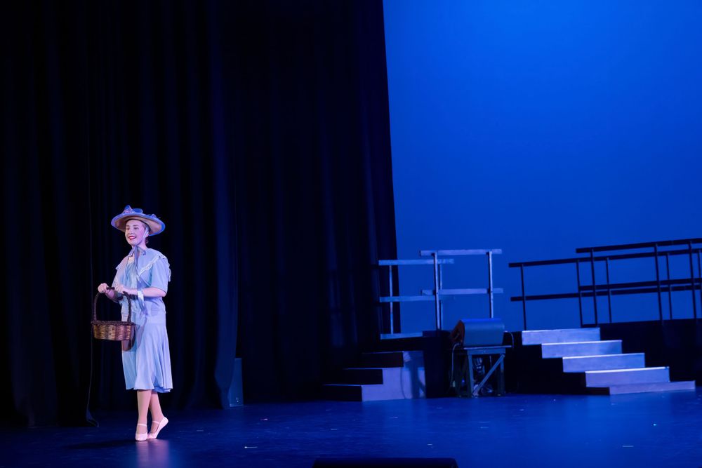 Solo performer stands on darkened stage with dramatic blue lighting and stair set piece.