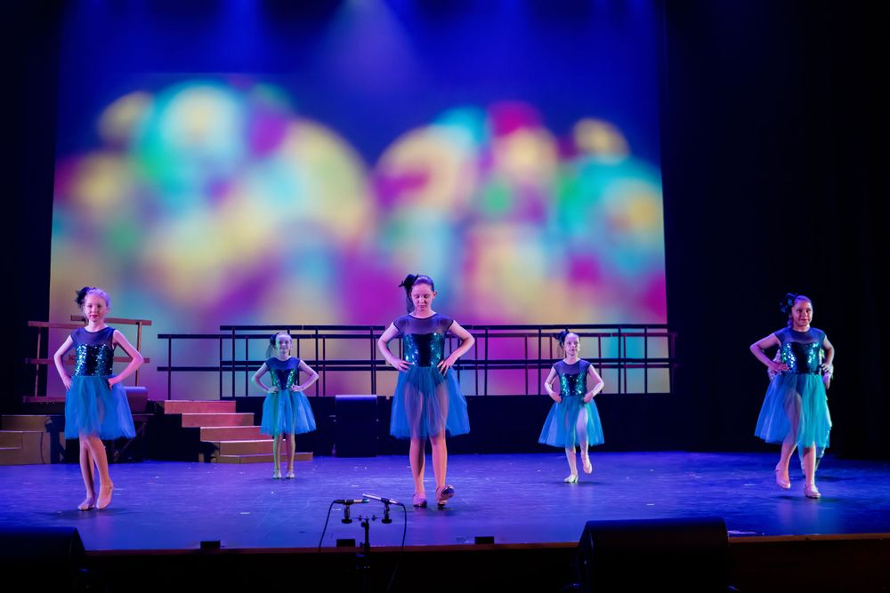 Dancers strike poses in blue dresses against a colorful bokeh backdrop on stage.