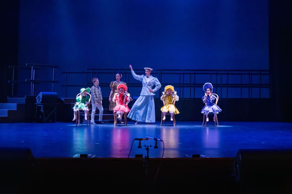 Dance performers in colorful costumes perform a theatrical number on a dimly lit stage with tiered platforms.