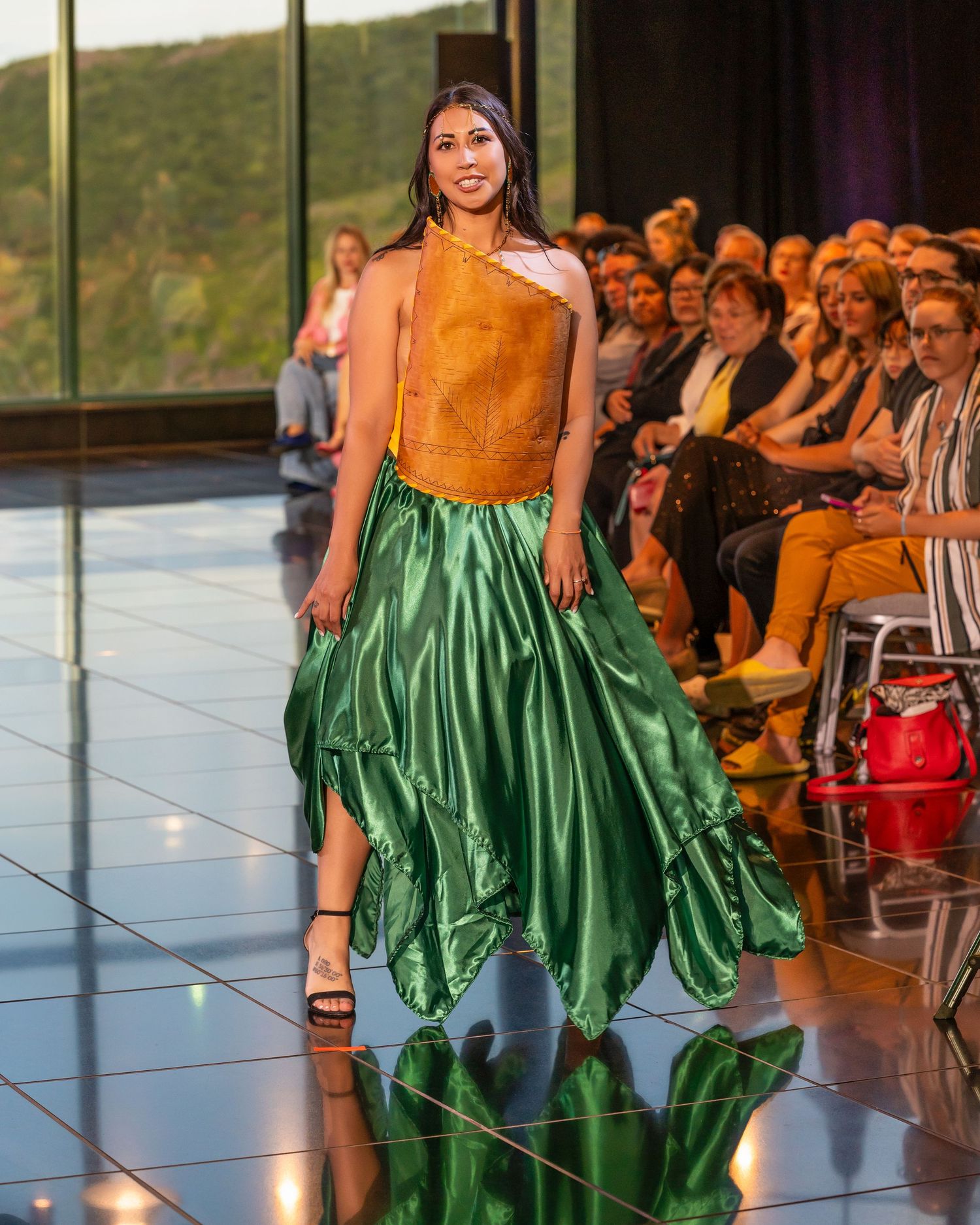 Model walks runway in orange sleeveless top and dramatic emerald green silk skirt at fashion show with audience seated.