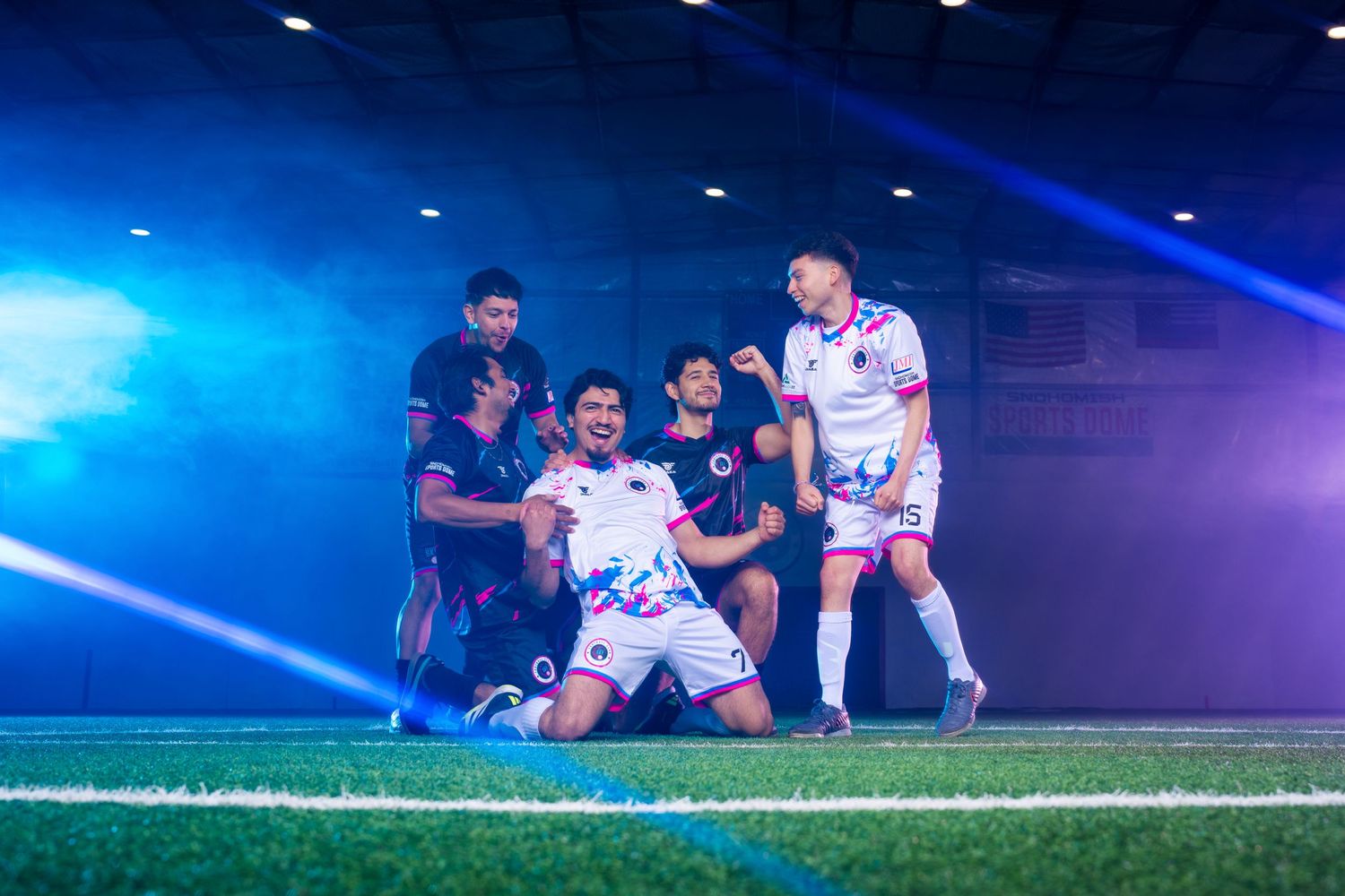 Soccer players in white and dark uniforms celebrating together on field with dramatic blue lighting and smoke effects.