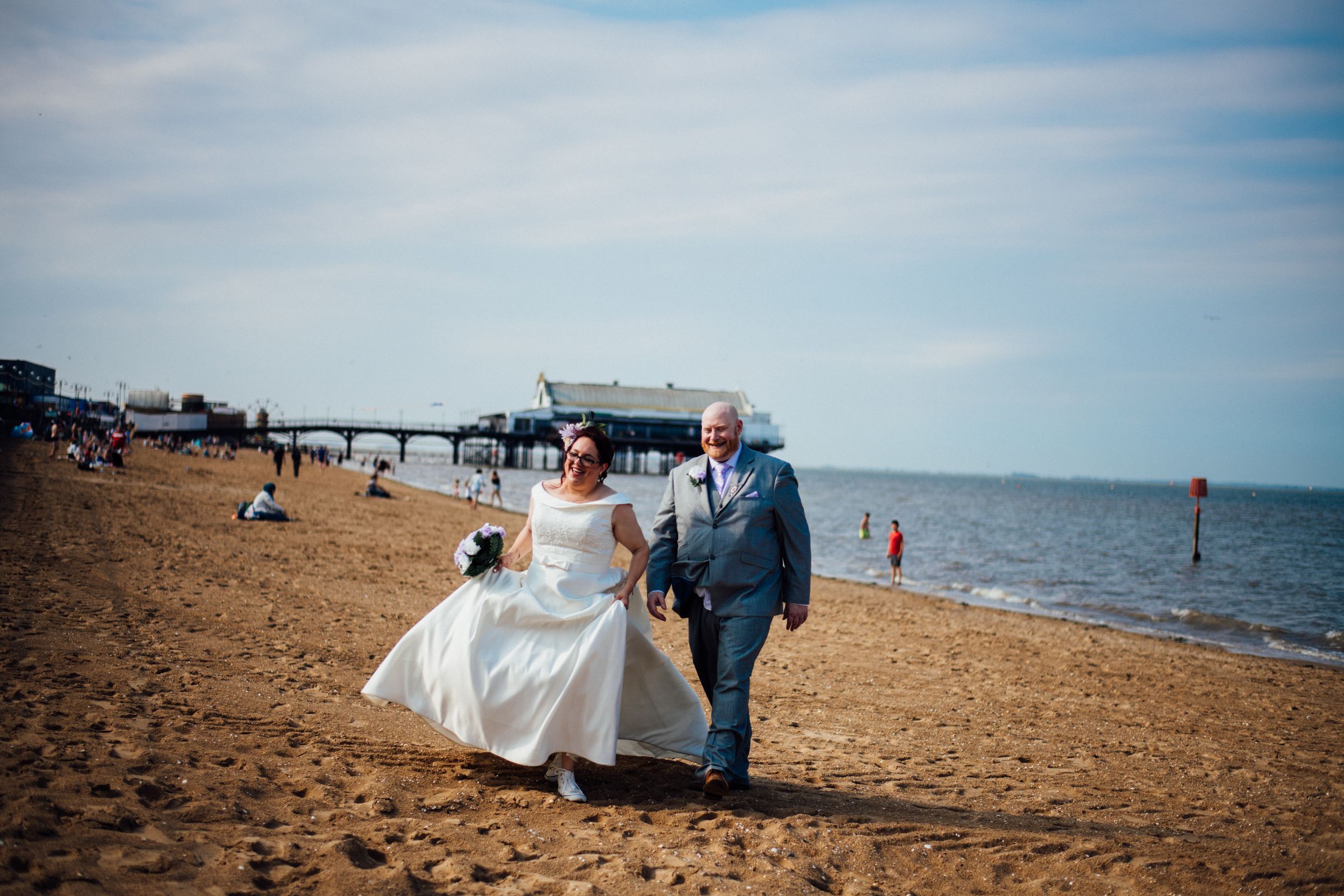 Wedding couple walking along sandy beach with pier in the distance.