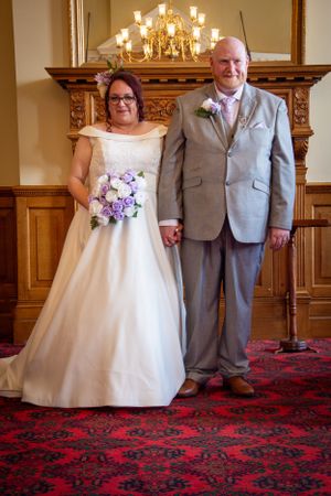 A wedding couple stands in front of an ornate wooden mantle with a chandelier in an elegant indoor setting.