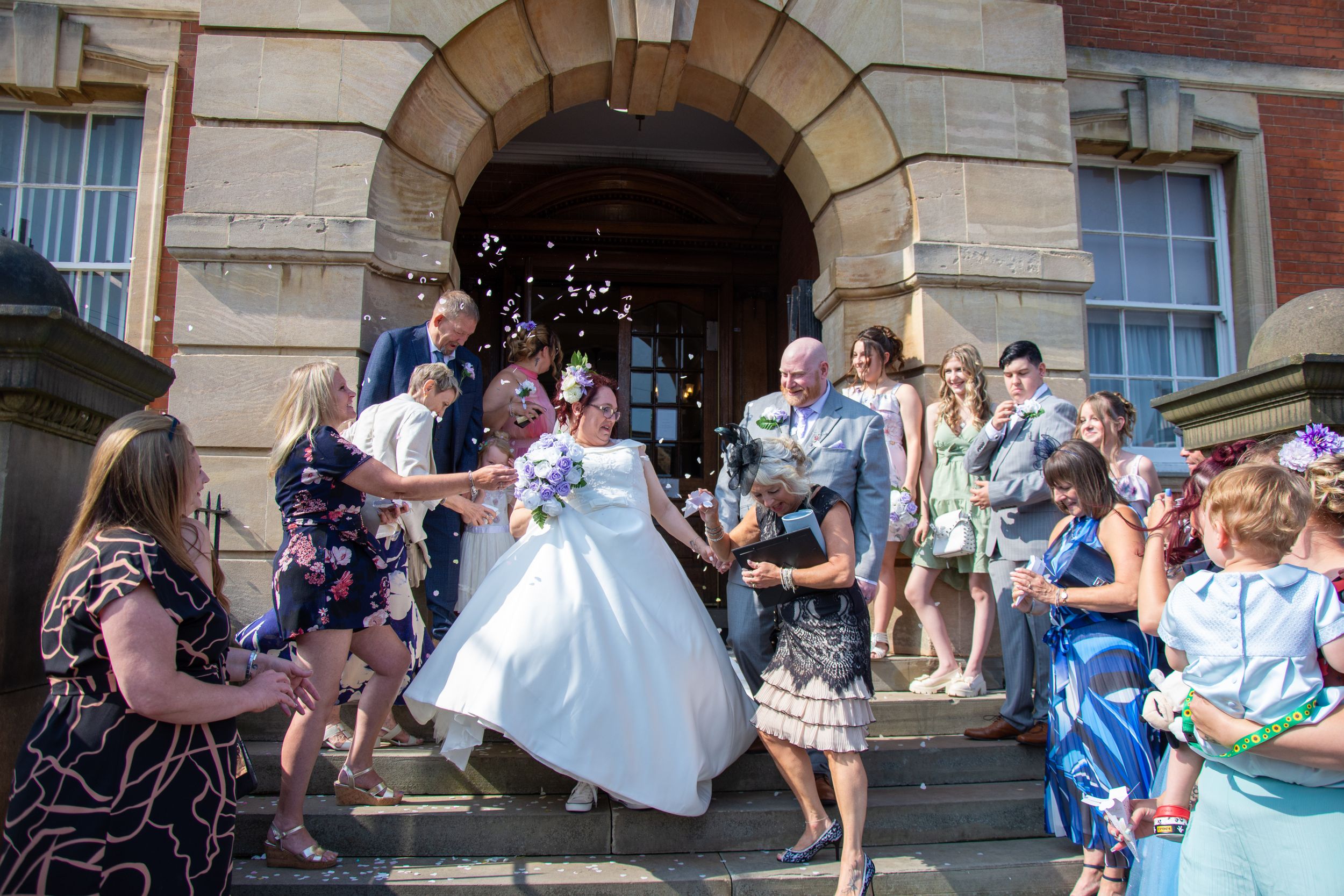 Wedding guests throw confetti on stone steps as celebrations happen outside a historic building.