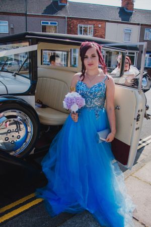 Woman in sparkly blue ball gown poses beside classic cream-colored car.